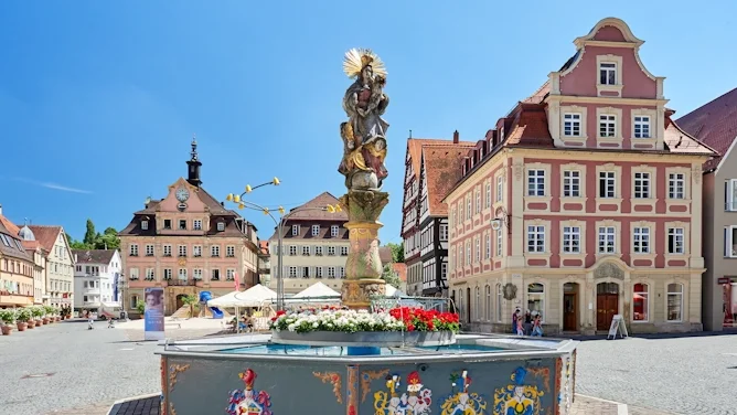 Blick auf einen historischen Marktplatz im Ostalbkreis mit Brunnen, klassischen Fassaden, roten Dächern, Fachwerkhäusern und blauem Himmel. Das Bild wird auf der Standortseite Ostalbkreis von GARANT Immobilien verwendet und steht für den regionalen Immobilienmarkt, attraktive Wohnlagen und Immobilienberatung in Aalen, Schwäbisch Gmünd, Ellwangen, Bopfingen, Lorch und Umgebung.
