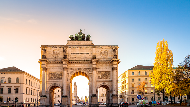 Blick auf das Siegestor in München mit historischer Architektur, umliegenden Gebäuden, Straße, blauem Himmel und warmem Licht. Das Bild wird auf der Standortseite München und Region von GARANT Immobilien verwendet und steht für den regionalen Immobilienmarkt, zentrale Wohnlagen und Immobilienberatung in München, Schwabing, Bogenhausen, Grünwald, Starnberg und Umgebung.
