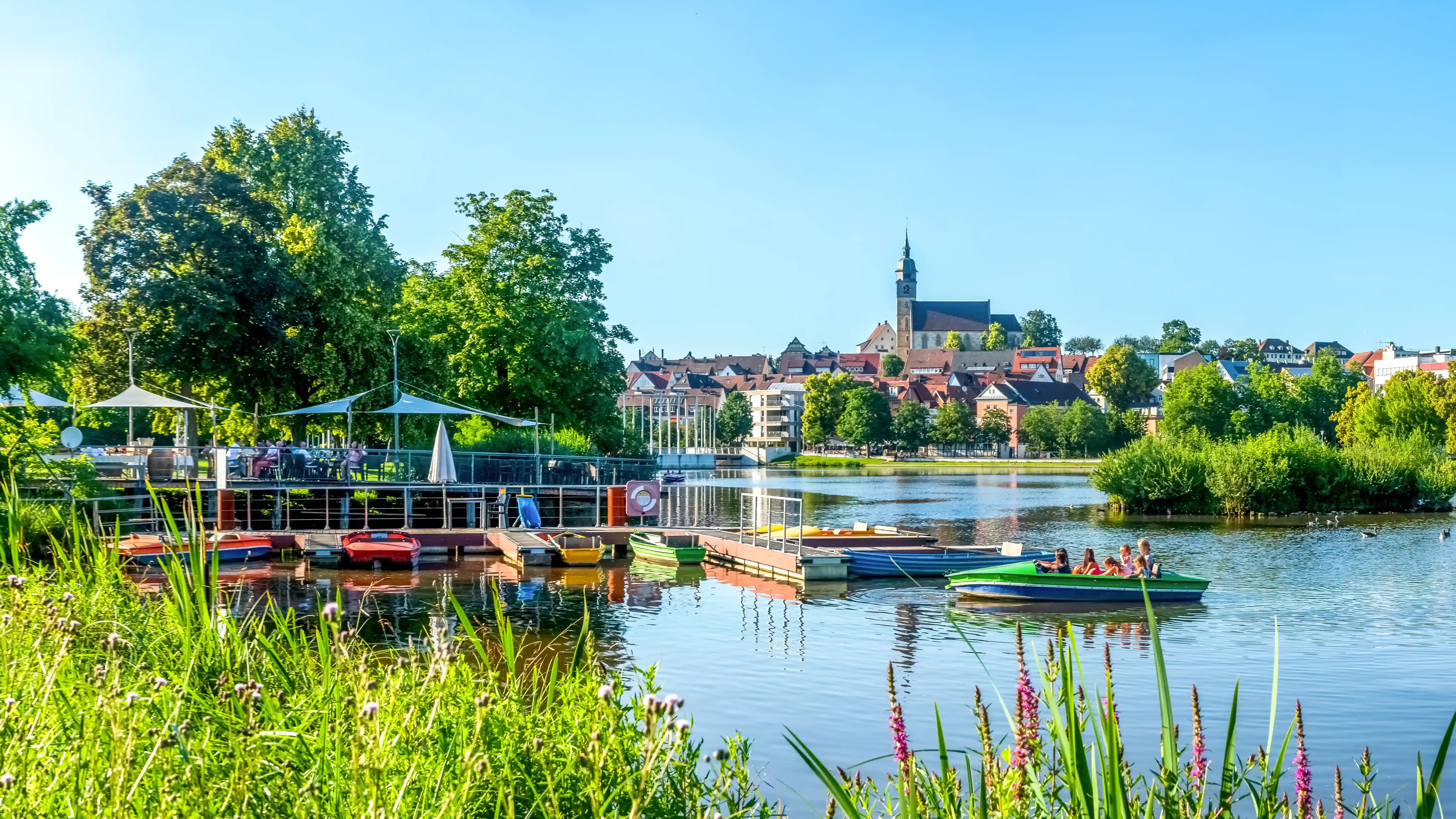 Blick auf den See in Böblingen mit grüner Uferlandschaft, Booten auf dem Wasser und der Stadt mit Kirchturm im Hintergrund. Das Bild wird als Header der Standortseite Böblingen von GARANT Immobilien verwendet und steht für regionale Immobilienberatung, lokale Marktkenntnis und persönliche Unterstützung beim Immobilienverkauf im Landkreis Böblingen.