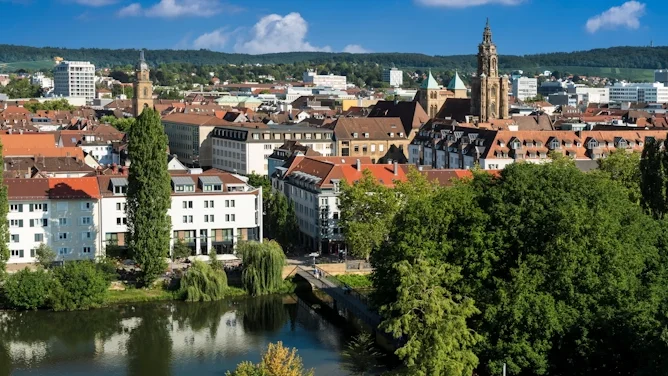 Blick auf Heilbronn mit Stadtgebäuden, Kirchtürmen, roten Dächern, grünen Uferbereichen und dem Neckar im Vordergrund. Das Bild wird auf der Standortseite Heilbronn von GARANT Immobilien verwendet und steht für den regionalen Immobilienmarkt, wirtschaftsstarke Wohnlagen und Immobilienberatung in Heilbronn, dem Heilbronner Land und Hohenlohe.