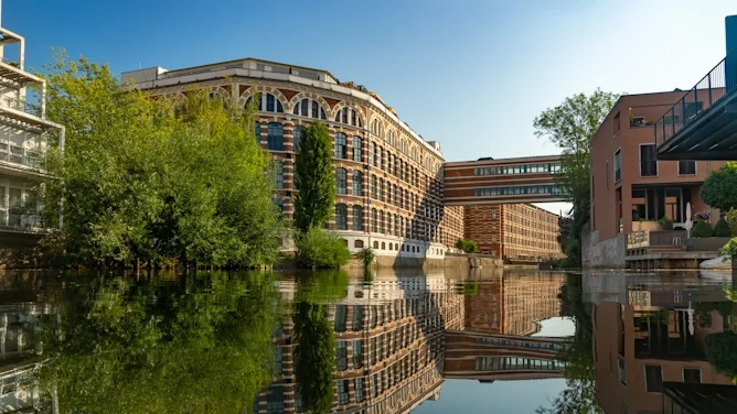 Blick auf eine moderne Wasserlage in Leipzig mit sanierten Gebäuden, Industriearchitektur, begrünten Uferbereichen und Spiegelung im Wasser. Das Bild wird auf der Standortseite Leipzig von GARANT Immobilien verwendet und steht für den regionalen Immobilienmarkt, urbane Wohnlagen, Kapitalanlagen und Immobilienberatung in Leipzig, Markkleeberg, Schkeuditz, Taucha und Umgebung.