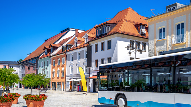 Blick auf eine Straße in der Deggendorfer Innenstadt mit farbigen Gebäuden, roten Dächern, blauem Himmel und einem Bus im Vordergrund. Das Bild wird auf der Standortseite Deggendorf von GARANT Immobilien verwendet und steht für den regionalen Immobilienmarkt, städtische Wohnlagen und Immobilienberatung in Deggendorf und Umgebung.