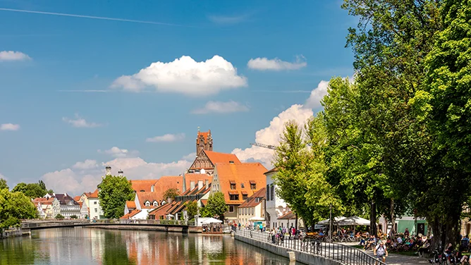 Blick auf Landshut mit Flussufer, historischen Gebäuden, roten Dächern, viel Grün, Spazierweg und blauem Himmel. Das Bild wird auf der Standortseite Landshut und Umgebung von GARANT Immobilien verwendet und steht für den regionalen Immobilienmarkt, attraktive Wohnlagen und Immobilienberatung in Landshut, Ergolding, Altdorf, Eching, Kumhausen und Umgebung.