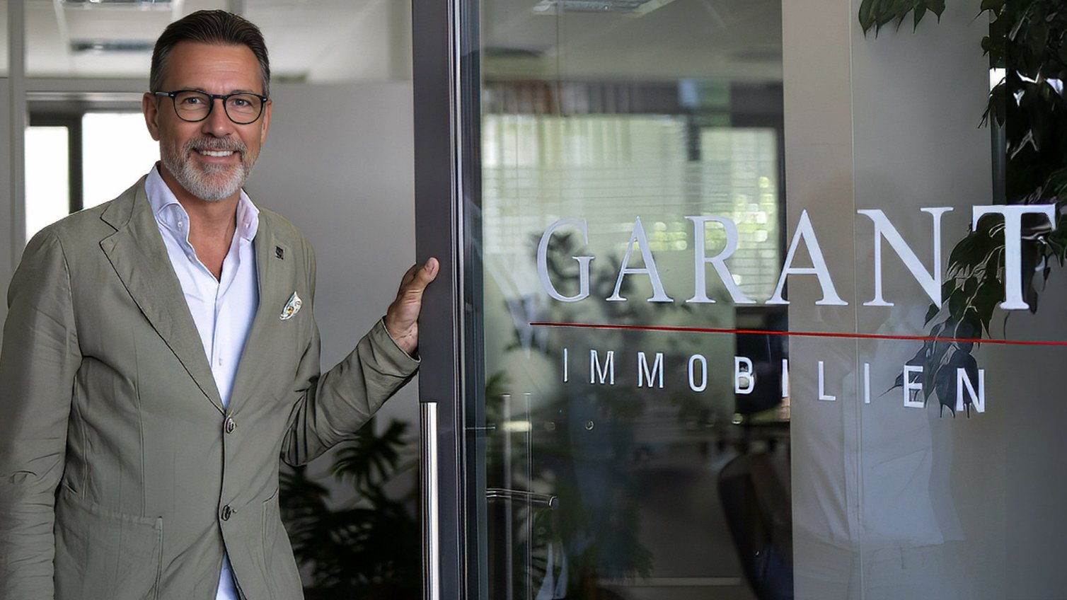 Ein Mitarbeiter steht lächelnd am Eingang eines GARANT Immobilien Büros und hält die Glastür geöffnet. Auf der Tür ist das GARANT Immobilien Logo zu sehen. Das Bild wird als Header der Über uns Seite verwendet und steht für Erfahrung, persönliche Beratung und regionale Stärke.