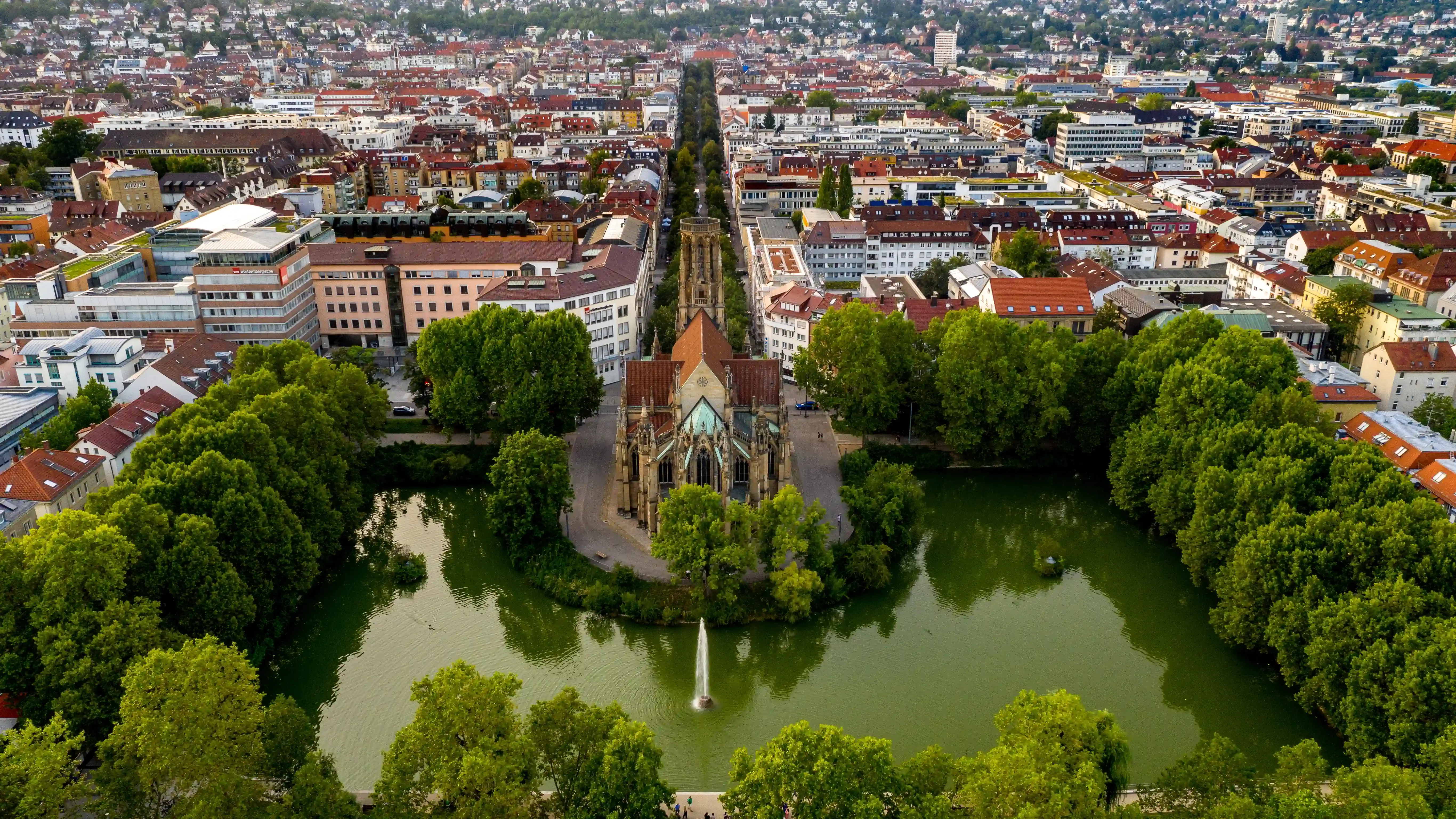 Luftaufnahme von Stuttgart mit dichter Stadtbebauung, grünen Parkflächen, See, Kirche und umliegenden Wohnquartieren. Das Bild wird auf der Standortseite Stuttgart von GARANT Immobilien verwendet und steht für den regionalen Immobilienmarkt, zentrale Wohnlagen und Immobilienberatung in Stuttgart, Stuttgart-West, Stuttgart-Ost, Vaihingen, Killesberg und Umgebung.