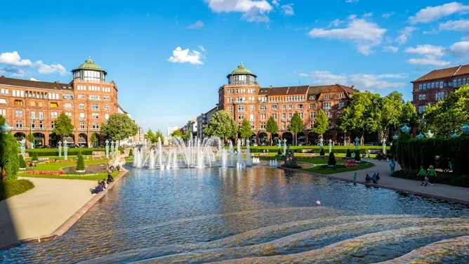 Blick auf Mannheim mit historischer Parkanlage, Wasserbecken, Brunnen, Backsteingebäuden, grünen Bäumen und blauem Himmel. Das Bild wird auf der Standortseite Mannheim von GARANT Immobilien verwendet und steht für den regionalen Immobilienmarkt, zentrale Wohnlagen und Immobilienberatung in Mannheim, Ludwigshafen, Viernheim, Heddesheim und Umgebung.