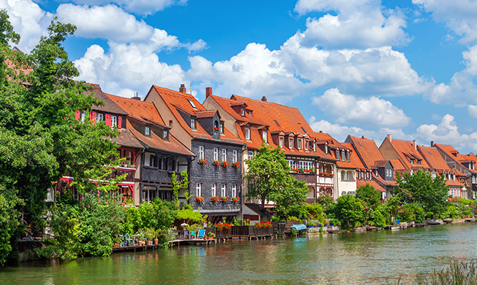 Blick auf die historische Altstadt von Bamberg mit Fachwerkhäusern, roten Dächern, grüner Uferbepflanzung und Fluss im Vordergrund. Das Bild wird auf der Standortseite Bamberg von GARANT Immobilien verwendet und steht für den regionalen Immobilienmarkt, die besondere Wohnqualität sowie Immobilienberatung in Bamberg und Umgebung.