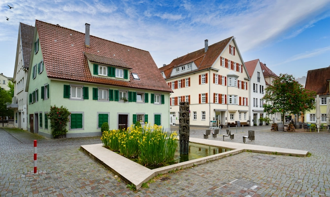 Blick auf einen historischen Platz in Göppingen mit hellen Gebäuden, roten Dächern, grünen Fensterläden, Brunnenanlage und gepflasterter Umgebung. Das Bild wird auf der Standortseite Göppingen von GARANT Immobilien verwendet und steht für den regionalen Immobilienmarkt, familienfreundliche Wohnlagen und Immobilienberatung in Göppingen, Uhingen, Eislingen, Süßen und Umgebung.