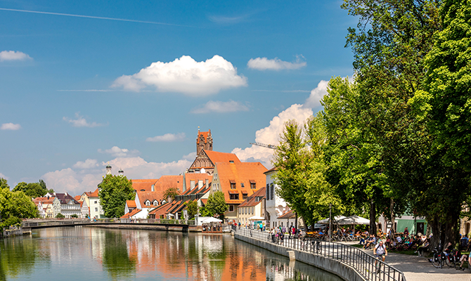 Blick auf Landshut mit Flussufer, historischen Gebäuden, roten Dächern, viel Grün, Spazierweg und blauem Himmel. Das Bild wird auf der Standortseite Landshut und Umgebung von GARANT Immobilien verwendet und steht für den regionalen Immobilienmarkt, attraktive Wohnlagen und Immobilienberatung in Landshut, Ergolding, Altdorf, Eching, Kumhausen und Umgebung.