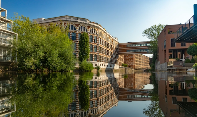 Blick auf eine moderne Wasserlage in Leipzig mit sanierten Gebäuden, Industriearchitektur, begrünten Uferbereichen und Spiegelung im Wasser. Das Bild wird auf der Standortseite Leipzig von GARANT Immobilien verwendet und steht für den regionalen Immobilienmarkt, urbane Wohnlagen, Kapitalanlagen und Immobilienberatung in Leipzig, Markkleeberg, Schkeuditz, Taucha und Umgebung.